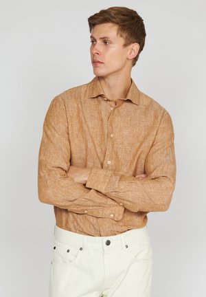 Light brown linen shirt with a button-down collar, long sleeves, and a textured fabric. Worn with white pants, arms crossed.