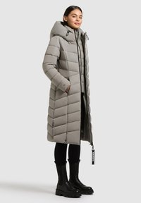 Langer, grauer Daunenmantel mit Chevron-Steppung, Kapuze und schwarzem Reißverschluss. Kombiniert mit schwarzen, klobigen Stiefeln.