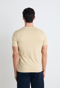 Beige bomull T-skjorte, kortermet, rund hals, slim fit, glatt tekstur, brukt med mørke bukser, sett fra baksiden.