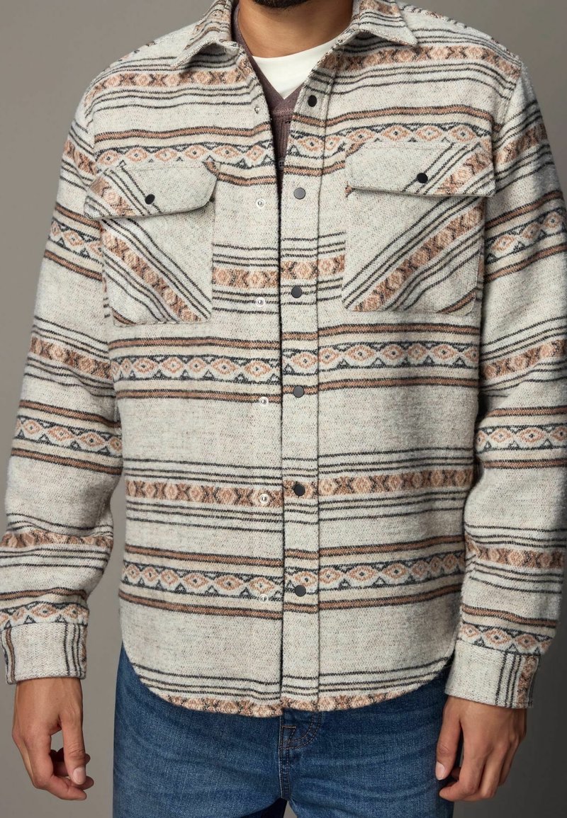 Homme portant une surchemise beige à motifs boutonnée avec deux poches poitrine et un jean bleu sur fond neutre.