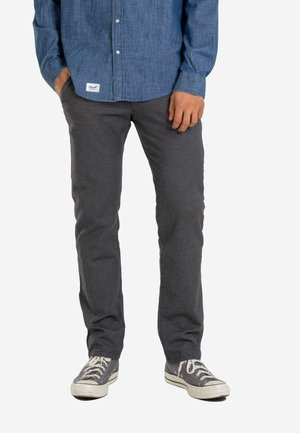 Man wearing dark gray textured pants, gray canvas sneakers with white laces, and a blue long-sleeve button-up shirt with a front tag.