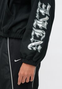 Άτομο που φοράει μαύρη κουκούλα με λευκή γοτθικού τύπου γραφή στο μανίκι και μαύρο αθλητικό παντελόνι Nike με λευκή ρίγα και λογότυπο, με το χέρι χαλαρά στο πλάι.