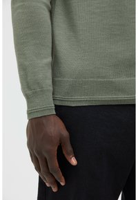 Olivgrüner, gestrickter Pullover mit gerippten Bündchen und Saum, kombiniert mit schwarzem Stoff. Die Textur wirkt glatt und weich.