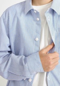 Niebieska i biała w paski koszula zapinana na guziki z kołnierzykiem, o gładkiej strukturze i widocznymi guzikami, noszona na białej koszulce.