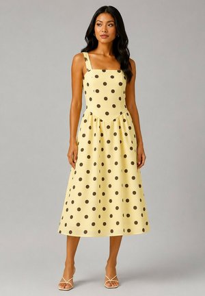 Femme portant une robe midi jaune sans manches à pois noirs avec un décolleté carré, debout dans une posture détendue sur un fond neutre.