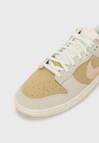 Baskets basses avec panneaux en daim beige et marron clair, lacets blancs et un logo Nike rose sur une semelle en caoutchouc blanche.