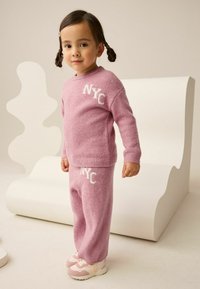 Rosa gestrickter Pullover und passende Hose mit "NYC" Stickerei. Gemütlicher Stoff, gerippte Bündchen und Bund, kombiniert mit hellrosa Sneakers.