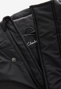 Czarna kurtka z gładką, strukturalną warstwą zewnętrzną, zatrzaskiem oraz widoczną metką Clarks w środku. Prosty, funkcjonalny design.