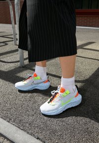 Vita sportskor med en fläckig sula, neonaccentuerad i gult och orange, och svarta snörningar. Bärs med vita strumpor och en svart randig kjol.