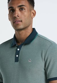 Groene en marineblauwe polo met een gestructureerd patroon, drie knopen en een klein pinguïnlogo op de linker borst.