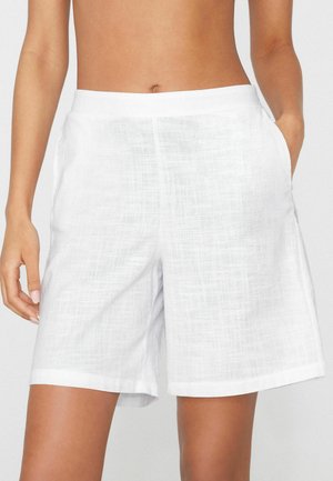 Shorts en lin blanc taille haute avec poches latérales portés par une personne avec le ventre et les bras nus visibles.
