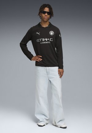 Junger Mann trägt ein schwarzes Manchester City Langarmtrikot, hellblaue weite Jeans, weiße Sneakers, dunkle Sonnenbrille und eine silberne Halskette.