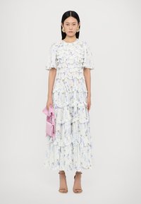 Lange, gelaagde jurk in lichtblauw met een witte bloemenprint, met gerimpelde lagen en korte mouwen. Gecombineerd met een roze clutch en beige hakken.