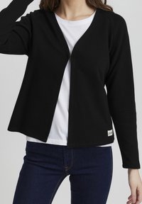 Černý kardigan s texturovanou tkaninou, otevřeným předním designem a dlouhými rukávy, spárovaný s bílým tričkem a tmavými džíny.