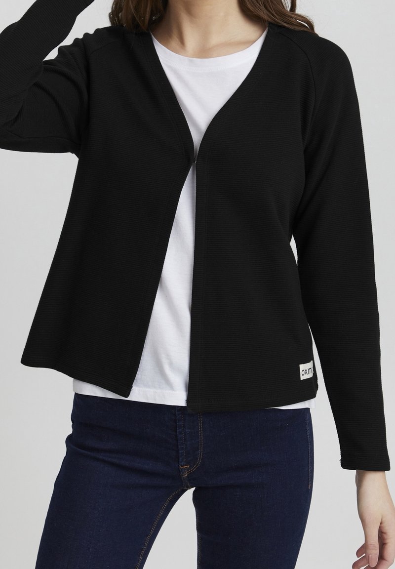 Černý kardigan s texturovanou tkaninou, otevřeným předním designem a dlouhými rukávy, spárovaný s bílým tričkem a tmavými džíny.