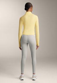 Dlouhá žlutá halenka s vysokým límcem, kombinovaná se světle šedými legínami. Outfit má přiléhavý střih a hladkou texturu.