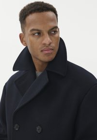 Cappotto navy doppiopetto con grande colletto, dotato di bottoni neri, realizzato in un tessuto spesso e textured. Sotto indossa un maglione grigio a maglia.