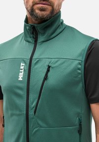 Grüne ärmellose Weste mit hohem Kragen, ausgestattet mit einem durchgehenden Reißverschluss und mehreren Reißverschlusstaschen. Strukturiertes Material mit schwarzen Akzenten und Branding.