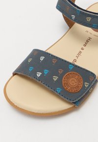 Grå skinnsandaler med et mønstret strop som har fargede diamantformer, en brun sirkulær logo som dekorasjon, og en beige såle.