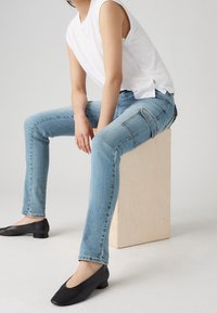 Jeans de denim azul claro con corte slim, que cuentan con bolsillos laterales y un ligero desvanecimiento. Combinados con zapatos negros de tacón bajo y una blusa sin mangas blanca.