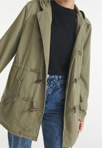 Kurtka w oliwkowym kolorze z kapturem, z zapięciem na guziki i ściągaczem w talii. Lekki materiał i widoczne kieszenie z przodu. Nosi się na czarnej bluzce.