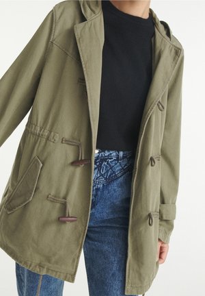 Olivgrön jacka med huva, toggelknäppningar och dragsko i midjan. Lättviktigt material och synliga framfickor. Bärs över en svart topp.