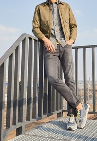 Uomo con giacca color khaki, camicia grigia e jeans appoggiato a un parapetto metallico all'aperto vicino ai binari del treno, indossando sneakers blu e bianche.