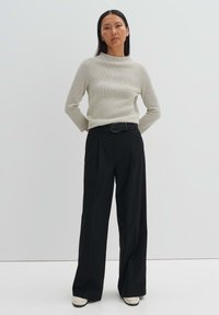 Gestrickter hellgrauer Oberteil mit hohem Kragen, kombiniert mit hoch sitzenden schwarzen Hosen. Das Outfit umfasst einen geflochtenen schwarzen Gürtel und cremefarbene Schuhe.