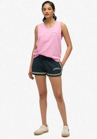 Jasnoróżowa bluzka bez rękawów z okrągłym dekoltem; czarne szorty z białymi paskami i logo "SUPERDRY"; neutralne kolorowe sneakersy.