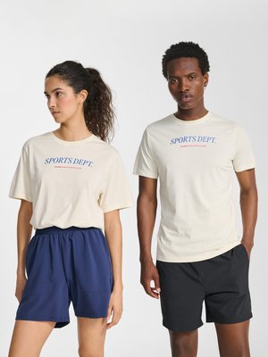 Zwei Models tragen cremefarbene T-Shirts mit der Aufschrift „SPORTS DEPT.“; das weibliche Model trägt marineblaue Shorts, das männliche Model trägt schwarze Shorts, beide stehen vor einem einfarbigen Hintergrund.