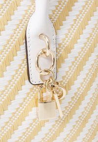 Woven bag z wzorem białym i żółtym, z białym paskiem i złotymi akcentami, w tym kłódką i ozdobnymi charmsami.