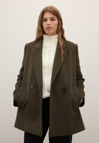 Abrigo de lana verde oliva con diseño de doble botonadura, solapas con muesca, bolsillos laterales y botones oscuros, sobre un suéter blanco texturizado.