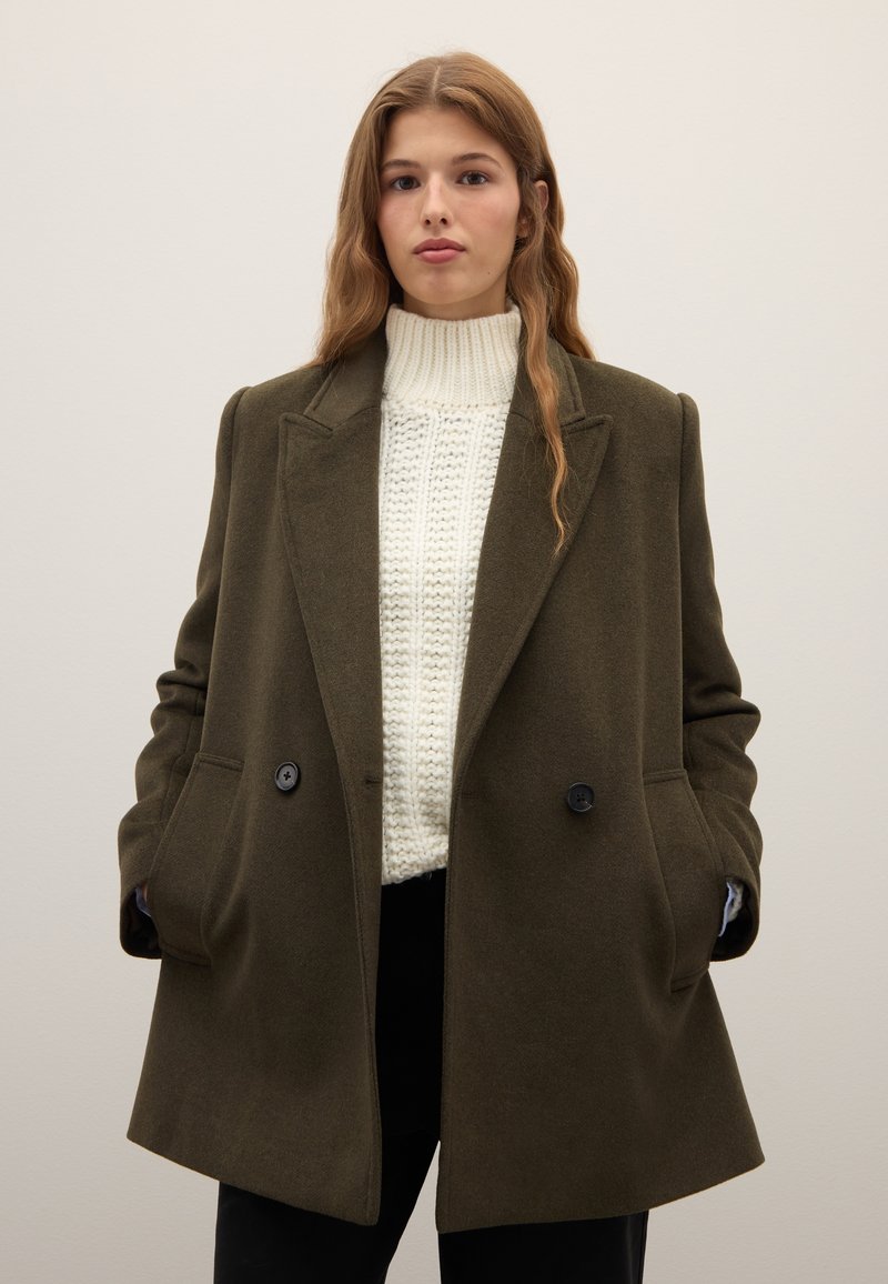 Abrigo de lana verde oliva con diseño de doble botonadura, solapas con muesca, bolsillos laterales y botones oscuros, sobre un suéter blanco texturizado.