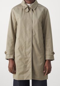 Manteau beige mi-long en coton ; il présente un col pointu, une fermeture boutonnée et des manches retroussées avec des boutons en accent.