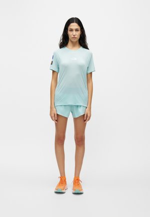 Donna in piedi con t-shirt sportiva celeste e pantaloncini con scarpe da corsa arancioni vivaci su sfondo bianco uniforme.