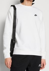 Sudadera blanca con cuello redondo, puños y dobladillo acanalados. Logo negro de Nike en el pecho. Bandolera negra con tirante llevada en el hombro.