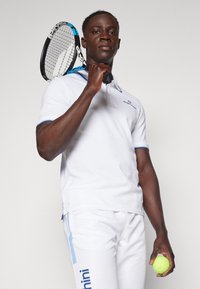 Polo bianco con accenti blu, abbinato a pantaloni bianchi con una striscia blu. Porta una racchetta da tennis e una pallina da tennis gialla.
