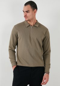 Maslinasto zelena dugosleeve polo majica s ovratnikom na zatvarač. Ima teksturiranu tkaninu i uski kroj, uparen s crnim hlačama.