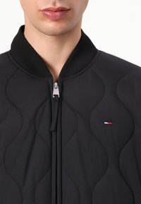 Black quilted jacket with a high collar, front zipper, and small embroidered logo featuring red, white, and blue accents on the chest.