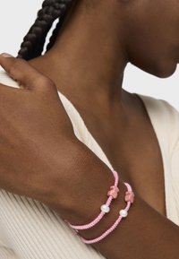 Due bracciali intrecciati rosa con accenti testurizzati e una perla bianca, indossati su un polso di carnagione scura contro una maglietta chiara.