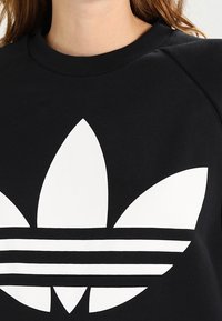 Černá mikina s velkým bílým logem trefoil. Obsahuje raglánové rukávy, kulatý výstřih a hladkou bavlněnou texturu.