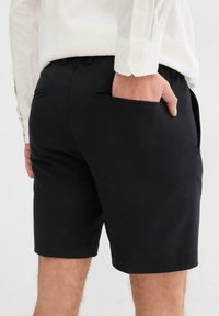 Pantaloni corti neri con una finitura in tessuto morbido, dotati di tasche laterali e una vestibilità sartoriale, abbinati a una camicia a bottone di colore chiaro.