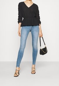 Černá texturovaná blůza s výstřihem do V a 3/4 rukávy, doplněná světle modrými skinny džínami. Modelka drží malou černou kabelku s řetízkem.