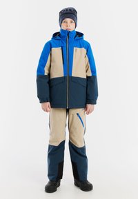 Niebieska i beżowa wodoodporna kurtka z kapturem i zapinanymi kieszeniami, zestawiona z pasującymi spodniami i czarnymi wodoodpornymi butami.