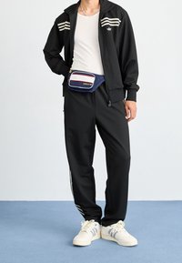 Černý teplákový komplet s bílými pruhy, bílý top bez rukávů, tmavě modrá ledvinka s červenými a bílými akcenty a krémové tenisky s šedými pruhy.