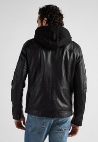 Zwarte leren jas met een gestructureerde afwerking, voorzien van een afneembare zwarte capuchon en schuine naden, gecombineerd met lichtblauwe jeans.