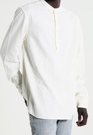 Chemise blanche à manches longues en lin avec col mandarin et patte de boutonnage portée par une personne portant un jean bleu.