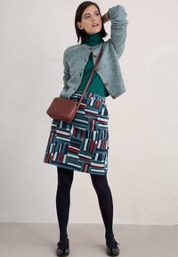 Grå stickad kofta, grön polotröja, mönstrad kjol med blå, röda och vita former. Svarta strumpbyxor och skinande svarta skor. Brun väska.