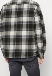 Vadderad flanellskjorta i svart, grått och vitt rutmönster. Har knäppning vid ärmsluten och rundad nederkant. Stylad med mörkgrå jeans.