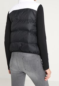 Svart och vit pufferväst med hög krage, kviltad design och en snygg finish. Bärs över en långärmad svart topp och grå jeans.